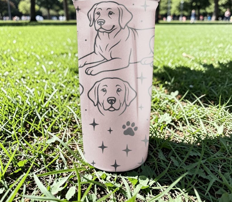 以下が含まれることがあります： ラブラドールレトリバーのイラストが描かれた、薄いピンク色のタンブラー。犬の足跡、星、2つの犬の肖像画が含まれています。円筒形のタンブラーは金属製のように見えます。