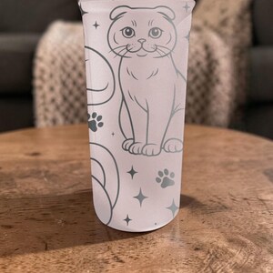 May include: A light pink tumbler featuring a gray illustration of a cat, paw prints, and stars. The cat is sitting upright with its ears folded down. The tumbler is cylindrical and appears to be made of a translucent material.