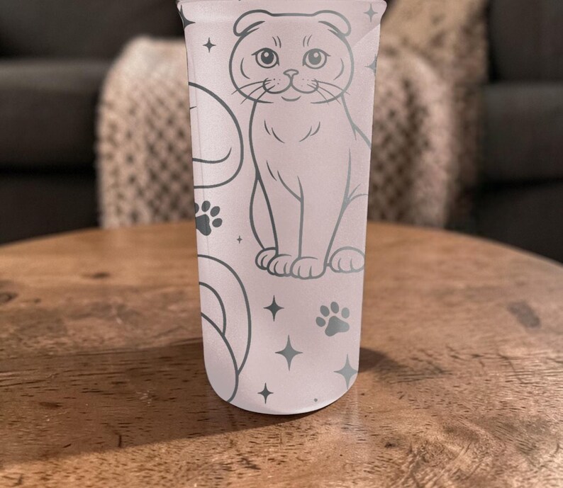 May include: A light pink tumbler featuring a gray illustration of a cat, paw prints, and stars. The cat is sitting upright with its ears folded down. The tumbler is cylindrical and appears to be made of a translucent material.