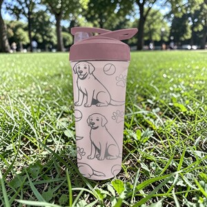 May include: A pink water bottle with a flip-top lid, featuring a repeating pattern of dog illustrations, paw prints, and tennis balls. The bottle is made of clear plastic and is suitable for carrying liquids. The lid is a darker shade of pink.