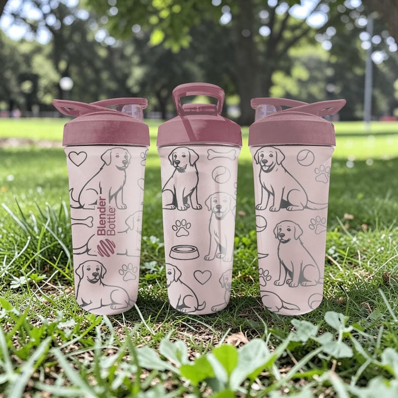 May include: Three pink Blender Bottles with maroon lids and handles, each decorated with a repeating pattern of dog illustrations, paw prints, and hearts. The bottles are on green grass.