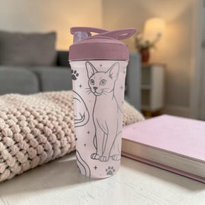 Puede incluir: Una botella de agua rosa con tapa abatible. La botella presenta una ilustraci&oacute;n de gato con huellas de patas y estrellas. La tapa y la parte superior de la botella son de un tono rosa m&aacute;s oscuro. La botella est&aacute; sobre una superficie blanca.