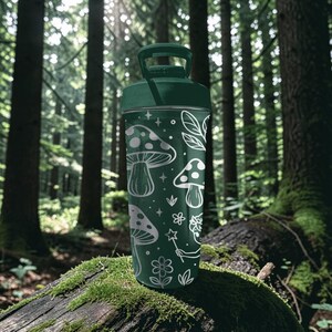 Puede incluir: Una botella de agua verde con tapa y asa a juego. La botella est&aacute; decorada con ilustraciones blancas de setas, estrellas, flores y hojas. La botella es cil&iacute;ndrica y parece estar hecha de metal o material similar.