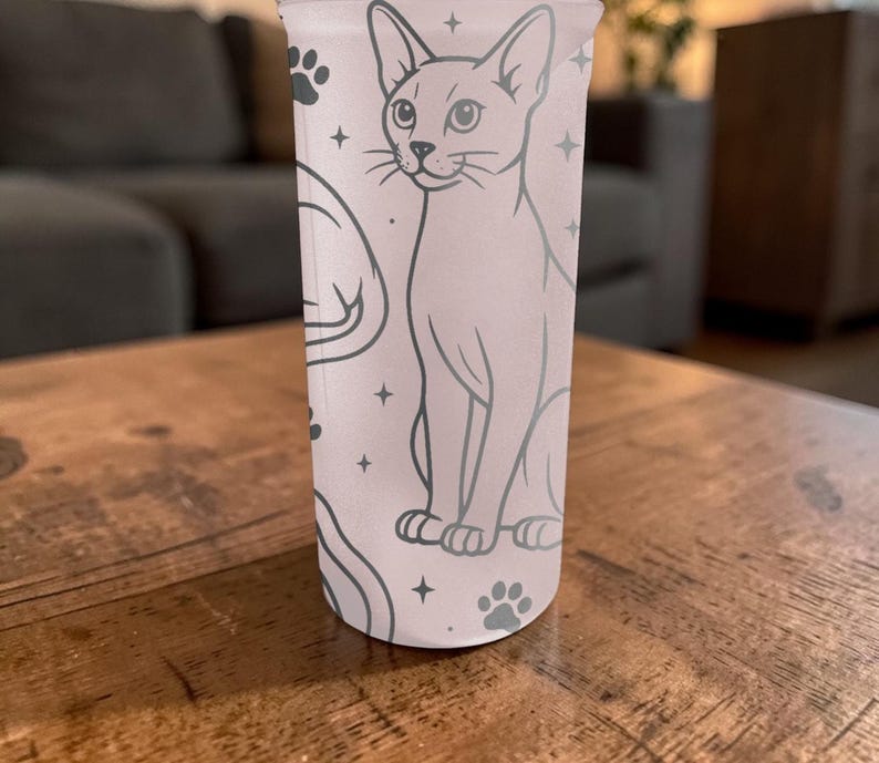 Puede incluir: Un vaso alto de color rosa claro con una ilustraci&oacute;n detallada de un gato, huellas de patas y estrellas. El gato est&aacute; representado en posici&oacute;n sentada con un contorno detallado. El dise&ntilde;o se repite alrededor del vaso.