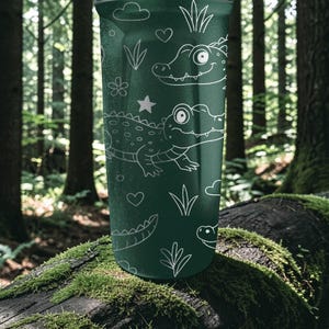 Peut inclure: Un gobelet vert fonc&eacute; avec des dessins au trait blancs de deux alligators de dessin anim&eacute;, des c&oelig;urs, des &eacute;toiles, des nuages et des plantes. Le gobelet est cylindrique. L'arri&egrave;re-plan est une sc&egrave;ne de for&ecirc;t floue.