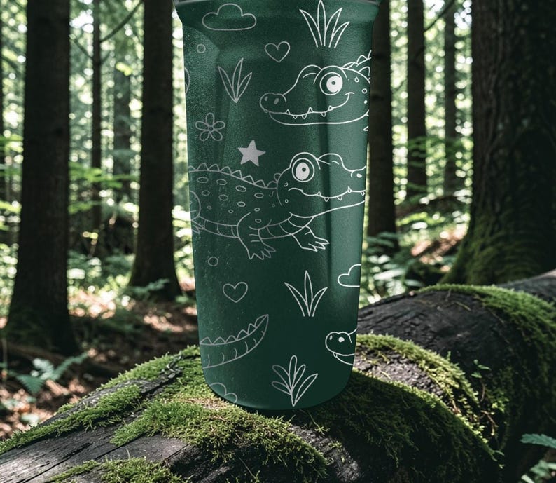 Peut inclure: Un gobelet vert fonc&eacute; avec des dessins au trait blancs de deux alligators de dessin anim&eacute;, des c&oelig;urs, des &eacute;toiles, des nuages et des plantes. Le gobelet est cylindrique. L'arri&egrave;re-plan est une sc&egrave;ne de for&ecirc;t floue.