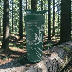 May include: A green water bottle with a black lid and a white graphic design. The design includes golf-related illustrations and the text "plzz not a BOGEY". The bottle is cylindrical and appears to be made of metal.