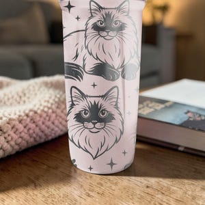 May include: A light pink tumbler featuring two black and white cat illustrations with star accents. The tumbler is cylindrical and appears to be made of metal or a similar material. The design is repeated on the tumbler's surface.