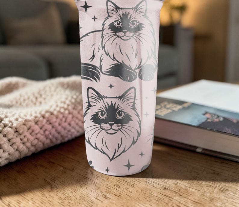 May include: A light pink tumbler featuring two black and white cat illustrations with star accents. The tumbler is cylindrical and appears to be made of metal or a similar material. The design is repeated on the tumbler's surface.