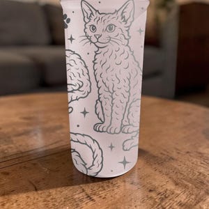 May include: A light pink tumbler with a cat illustration, brain, and rope design. The cat is sitting, and the design is in a light gray color. The tumbler has a straight shape and is made of a translucent material.