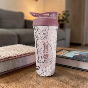 May include: A Blender Bottle with a light pink body and a dark pink lid. The bottle features a black and white illustration of a cat, paw prints, and stars. The Blender Bottle logo is also visible.