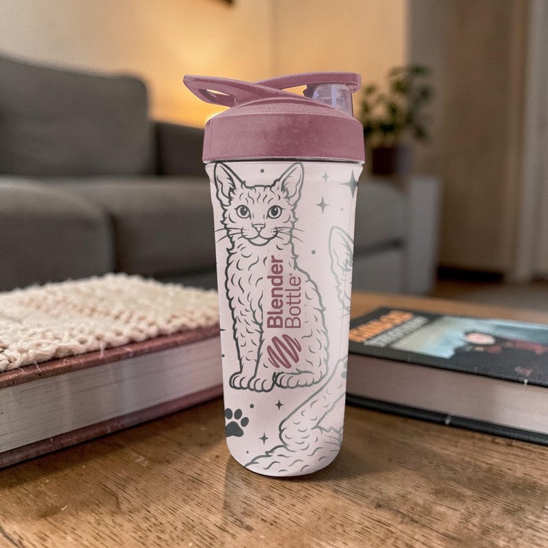 May include: A Blender Bottle with a light pink body and a dark pink lid. The bottle features a black and white illustration of a cat, paw prints, and stars. The Blender Bottle logo is also visible.