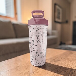 May include: A clear water bottle with a maroon lid and handle. The bottle is decorated with a repeating pattern of Scottish Fold cat illustrations and small star shapes. The bottle is sitting on a wooden table.