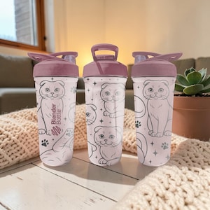 May include: Three Blender Bottles with a light purple lid and base. The bottles have a white background with a repeating pattern of a cat illustration, paw prints, and stars. The text "Blender Bottle" is printed on the side of one bottle.