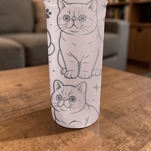 May include: A tall, clear tumbler with a repeating pattern of cartoon cats in light grey. The cats are drawn in a simple, outlined style, with details like whiskers and large eyes. The tumbler is set on a wooden surface.