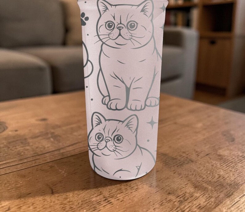 May include: A tall, clear tumbler with a repeating pattern of cartoon cats in light grey. The cats are drawn in a simple, outlined style, with details like whiskers and large eyes. The tumbler is set on a wooden surface.