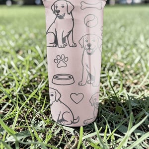 May include: A pink tumbler decorated with black line drawings of Labrador Retrievers, paw prints, a dog bowl, a bone, a ball, and hearts. The tumbler is cylindrical and appears to be made of metal or plastic.