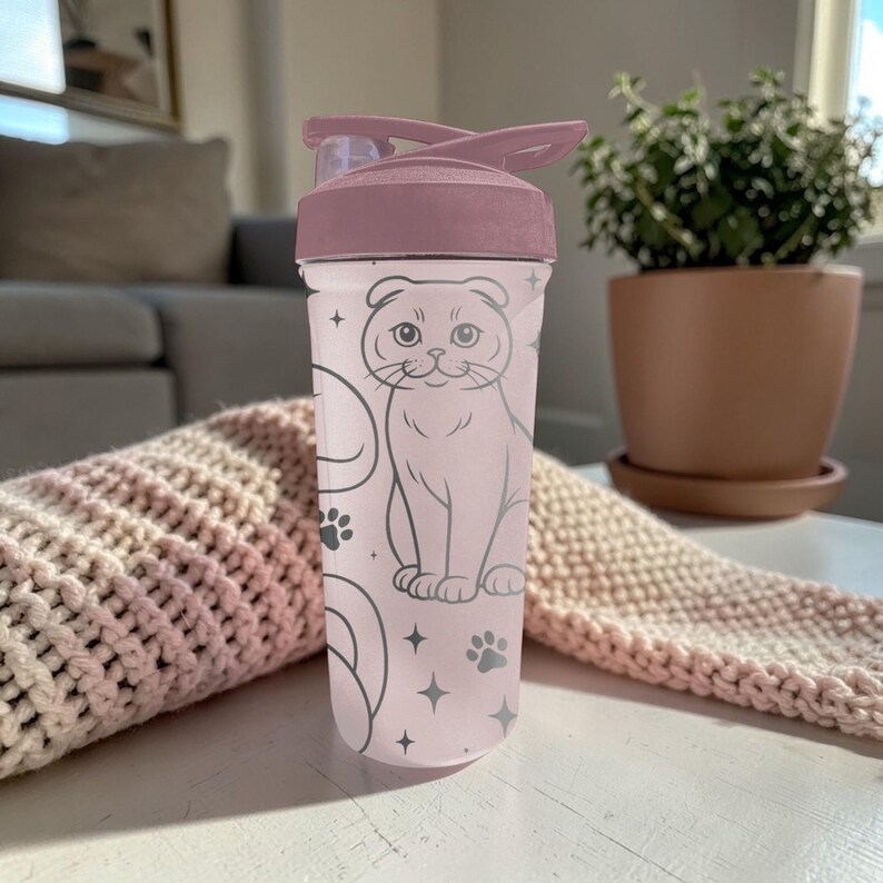 May include: A pink water bottle with a flip-top lid. The bottle features a gray illustration of a cat, paw prints, and stars. The lid and top portion of the bottle are a darker shade of pink. The bottle is sitting on a white surface.