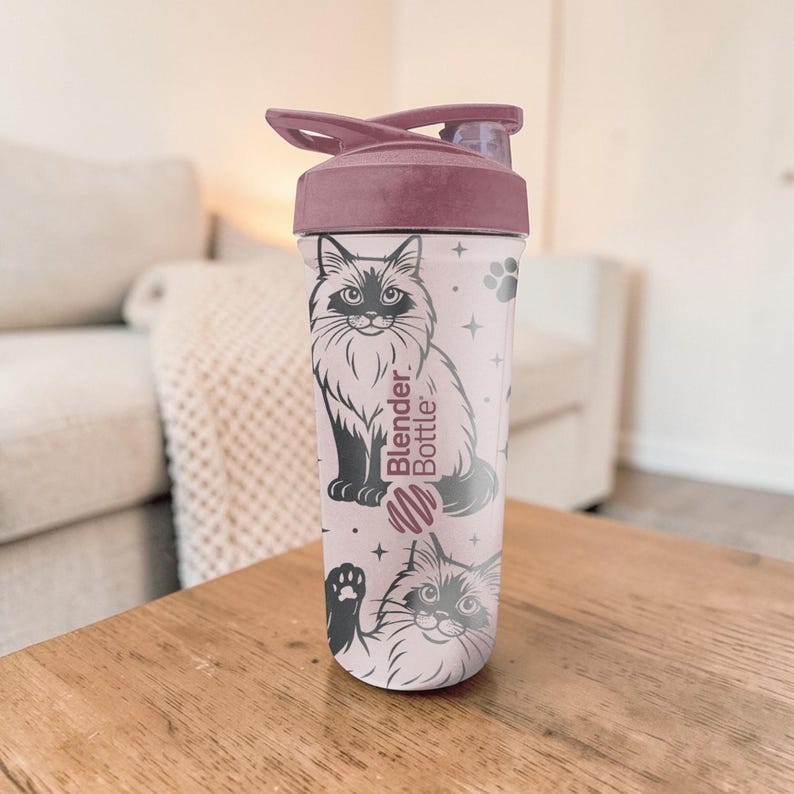 May include: A pink Blender Bottle with a dark pink lid. The bottle features a black and white illustration of a cat, paw prints, and stars. The words "Blender Bottle" are printed on the side.