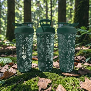 May include: Three green Blender Bottles with white lizard and leaf designs. Each bottle has a dark green lid and handle. The bottles are displayed outdoors on a bed of green moss and fallen leaves, with a forest background.