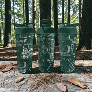 May include: Three green Blender Bottles with golf-themed designs. Each bottle features white illustrations of golf equipment, golfers, and text such as "Mulligan" and "plzz not a bogey". The bottles are on a wooden surface with fallen leaves, set against a blurred forest backdrop.