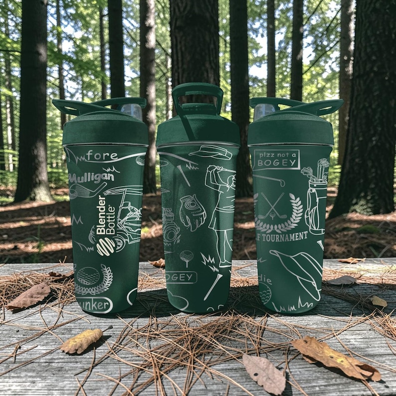 May include: Three green Blender Bottles with golf-themed designs. Each bottle features white illustrations of golf equipment, golfers, and text such as "Mulligan" and "plzz not a bogey". The bottles are on a wooden surface with fallen leaves, set against a blurred forest backdrop.