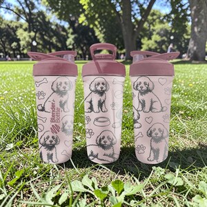May include: Three pink Blender Bottles with a dog-themed design. Each bottle features a repeating pattern of a puppy, paw prints, and bones. The lids and flip-top closures are a matching pink color. The text "Blender Bottle" is printed on the side of one bottle.