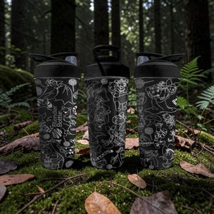 May include: Three black Blender Bottles with white cartoon character designs. The bottles are on moss and leaves in a forest. Each has a black lid with a flip-top opening. The text "Blender Bottle" is visible on one bottle.