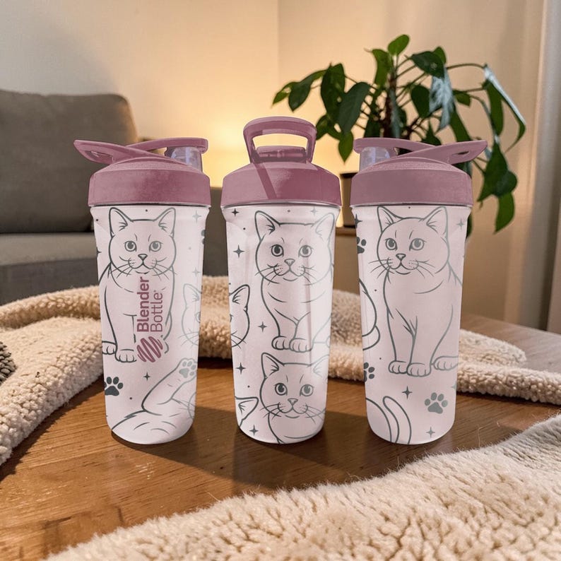 May include: Three Blender Bottle brand shaker cups with a cat design. The cups have a light pink lid and a clear body with a cat illustration. The text "Blender Bottle" is printed on the side of one cup.
