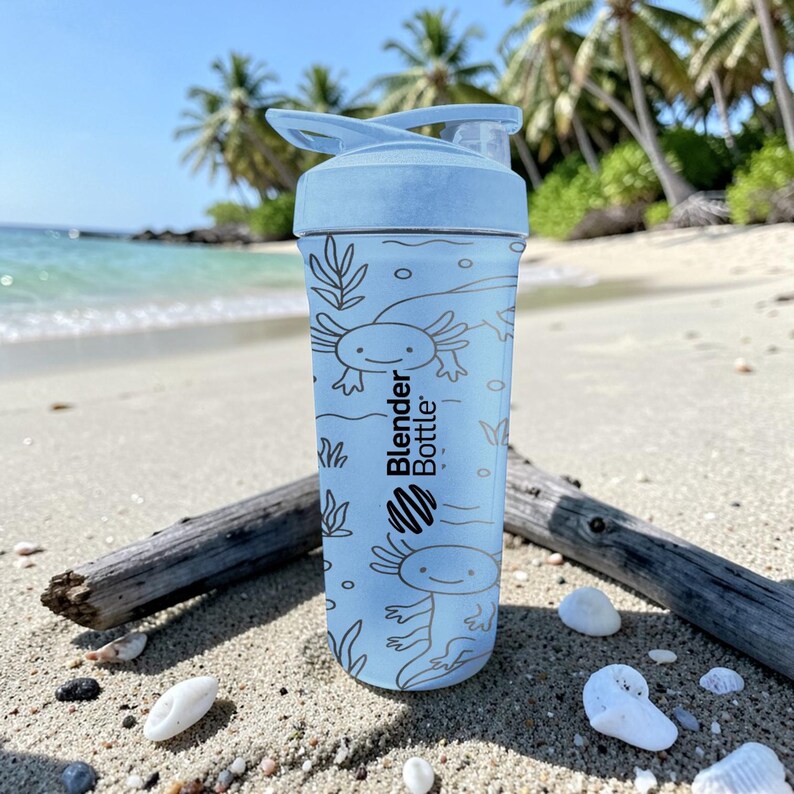 May include: A light blue Blender Bottle water bottle with a flip-top lid. The bottle features a repeating pattern of axolotl illustrations and the brand name. The bottle is sitting on a sandy beach with driftwood and seashells.