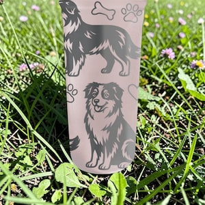 K&ouml;nnte beinhalten: Ein hellrosa Becher mit zwei Hundeillustrationen, Pfotenabdr&uuml;cken, einem Knochen und Herzen. Ein Hund steht, der andere sitzt. Der Becher ist von gr&uuml;nem Gras und kleinen lila Blumen umgeben.