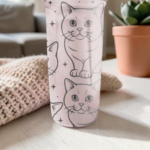 May include: A light pink tumbler featuring a repeating pattern of cat illustrations and star accents. The cats are drawn in a simple, cartoon style with black outlines. The tumbler is cylindrical and appears to be made of plastic.