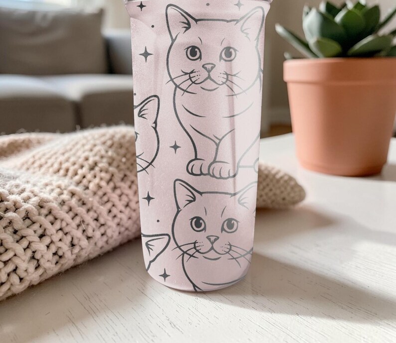 May include: A light pink tumbler featuring a repeating pattern of cat illustrations and star accents. The cats are drawn in a simple, cartoon style with black outlines. The tumbler is cylindrical and appears to be made of plastic.