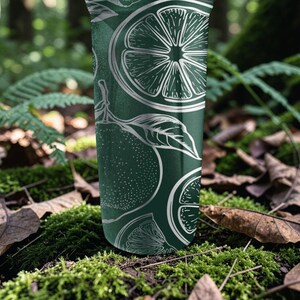 May include: A dark green tumbler with a white citrus fruit design. The design includes whole citrus fruits, slices, and leaves. The tumbler is cylindrical and appears to be made of metal. The background is a natural setting with green moss and fallen leaves.