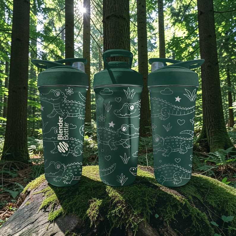 Peut inclure: Trois bouteilles Blender Bottle vertes avec des motifs d'alligators et de gribouillis blancs. Les bouteilles ont un couvercle rabattable et une poign&eacute;e. Elles sont pos&eacute;es sur une b&ucirc;che moussue dans une for&ecirc;t. Le logo Blender Bottle est visible.
