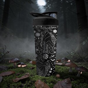 Pu&ograve; includere: Bottiglia Blender nera con un design floreale e pipistrello bianco. La bottiglia ha un coperchio nero e un beccuccio trasparente. La bottiglia si trova in un ambiente forestale con funghi e foglie.