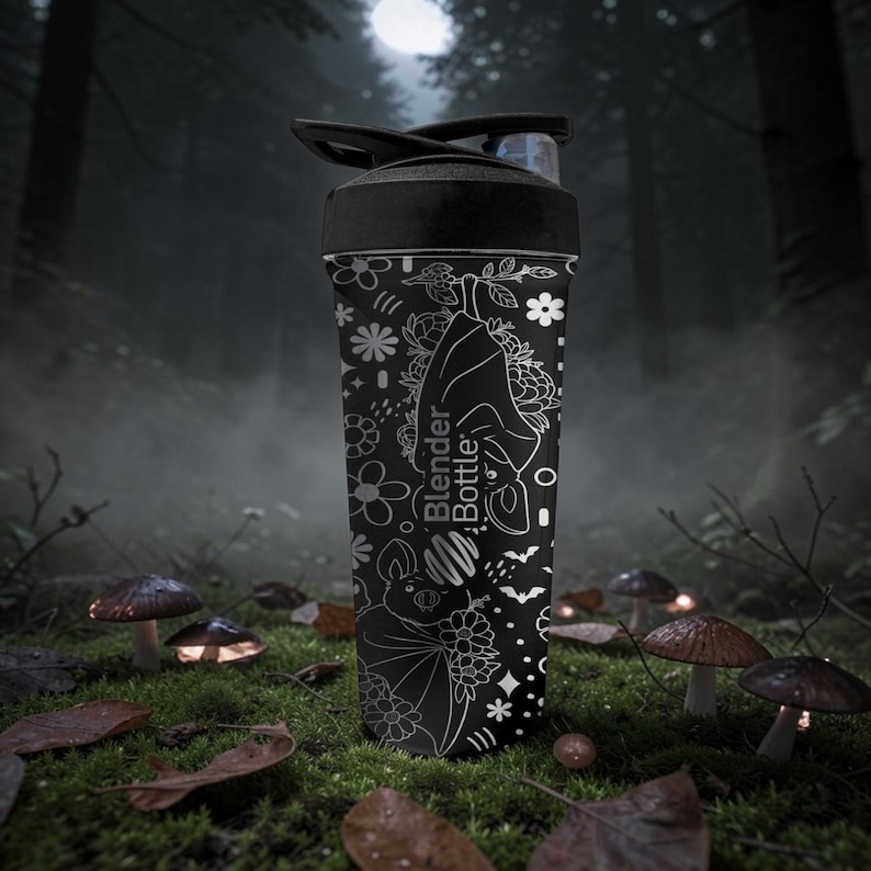 Pu&ograve; includere: Bottiglia Blender nera con un design floreale e pipistrello bianco. La bottiglia ha un coperchio nero e un beccuccio trasparente. La bottiglia si trova in un ambiente forestale con funghi e foglie.