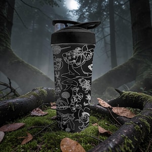 May include: Black water bottle with a black lid, decorated with white line art of cartoon characters and Halloween-themed illustrations. The bottle is cylindrical and made of plastic. The background is a forest scene with moss and fallen leaves.