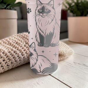 May include: A light pink tumbler featuring a gray illustration of a fluffy cat. The design includes paw prints and stars. The tumbler is cylindrical and appears to be made of a translucent material.
