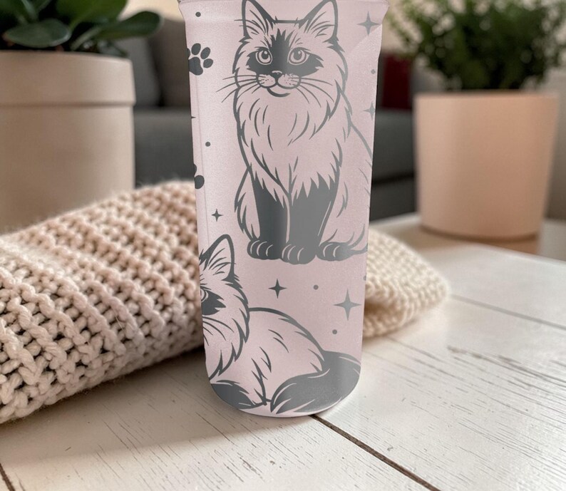 May include: A light pink tumbler featuring a gray illustration of a fluffy cat. The design includes paw prints and stars. The tumbler is cylindrical and appears to be made of a translucent material.