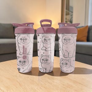 May include: Three white Blender Bottles with a light pink lid and handle. Each bottle features a repeating pattern of a cartoon cat and stars. One bottle has the text "Blender Bottle" printed on it.