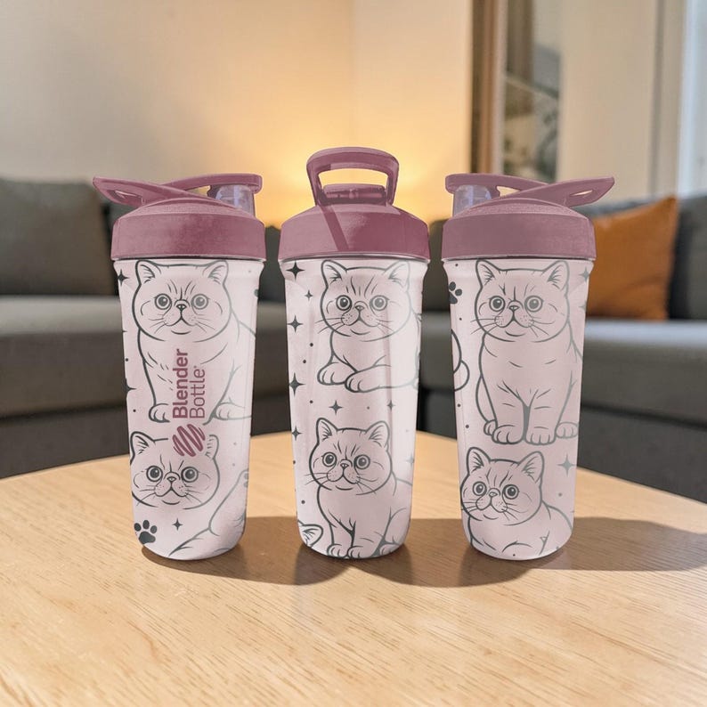 May include: Three white Blender Bottles with a light pink lid and handle. Each bottle features a repeating pattern of a cartoon cat and stars. One bottle has the text "Blender Bottle" printed on it.