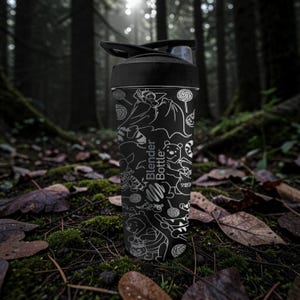 May include: Black Blender Bottle with a black lid, decorated with white line art of cartoon characters and sweets. The bottle has the words "Blender Bottle" printed on it. The background is a forest scene with moss and fallen leaves.