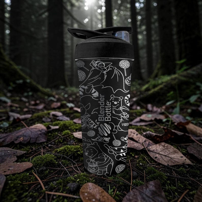 May include: Black Blender Bottle with a black lid, decorated with white line art of cartoon characters and sweets. The bottle has the words "Blender Bottle" printed on it. The background is a forest scene with moss and fallen leaves.