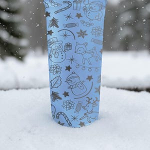 Puede incluir: Un vaso azul claro con un dise&ntilde;o de vacaciones de invierno. El dise&ntilde;o incluye mu&ntilde;ecos de nieve, renos, bastones de caramelo, estrellas y copos de nieve. El vaso est&aacute; en la nieve.