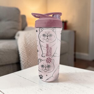 May include: A pink Blender Bottle with a matching lid. The bottle features a repeating pattern of a cartoon cat with stars and paw prints. The text "Blender Bottle" is printed on the side of the bottle.