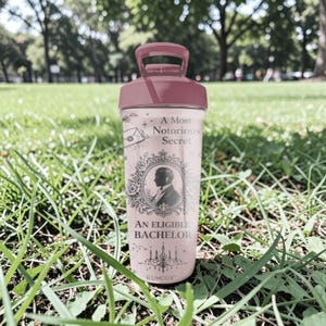 Pode incluir: Uma garrafa de &aacute;gua rosa e gren&aacute; com uma al&ccedil;a. A garrafa apresenta o texto "A Most Notorious Secret" e "An Eligible Bachelor" com a silhueta de um homem em uma moldura decorativa. A palavra "RUMOUR" est&aacute; na parte inferior.