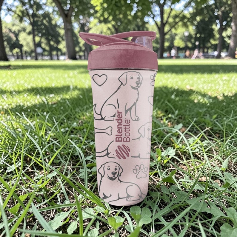 May include: A pink Blender Bottle with a maroon lid and flip-top. The bottle features a repeating pattern of dog illustrations, hearts, paw prints, and the Blender Bottle logo. The bottle is sitting on green grass.