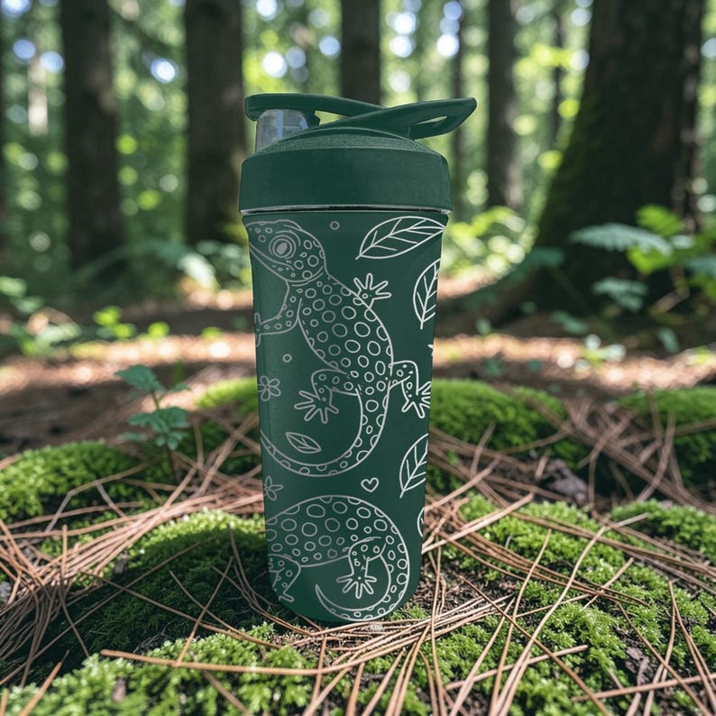 May include: A dark green water bottle with a white lizard and leaf pattern. The bottle has a flip-top lid and is set against a forest background. The bottle is approximately 9 inches tall.