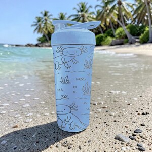 May include: A light blue water bottle featuring a cartoon axolotl design. The bottle has a flip-top lid and is decorated with axolotls, aquatic plants, and bubbles. The bottle is shown on a sandy beach with palm trees in the background.